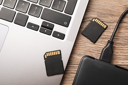 Hard disk drive with USB cable, SD memory cards and laptop on wooden table, flat lay