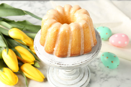 Delicious bundt cake, Easter eggs and tulips on white marble table, closeup