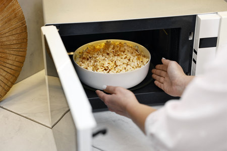 Woman taking bowl with popcorn out of microwave oven at white marble countertop in kitchen, closeupの写真素材