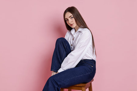 Beautiful young woman in stylish jeans sitting on stool against pink backgroundの写真素材