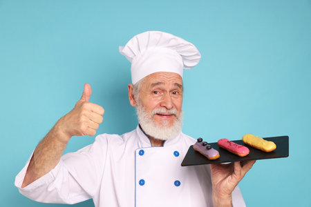 Professional pastry chef with delicious eclairs showing thumbs up on light blue backgroundの写真素材