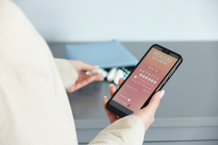 Menstruation. Woman holding smartphone with period calendar application and taking tampon indoors, closeupの写真素材