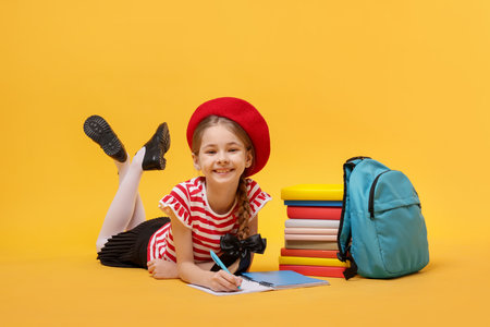 Schoolgirl with backpack and stack of books doing homework on yellow backgroundの写真素材