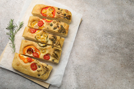 Cut delicious focaccia with flowers made of vegetables and rosemary on gray table, top view. Space...