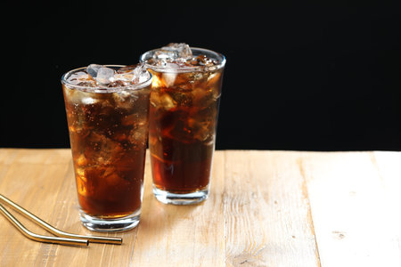 Refreshing cola with ice cubes in glasses and straws on wooden table against black background,...