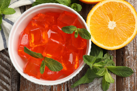 Delicious jelly cubes in bowl, mint and orange on wooden table, flat lay
