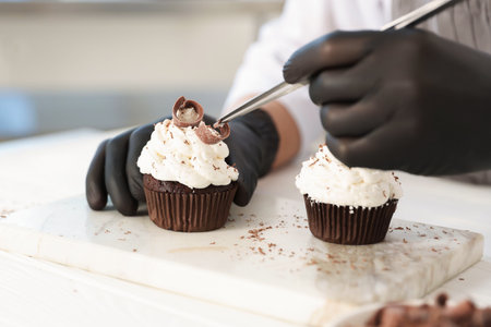 Confectioner decorating tasty cupcake with chocolate shaving at white wooden table in kitchen, closeupの写真素材