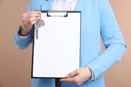 Real estate agent holding key with house shaped keychain and clipboard on beige background, closeupの写真素材