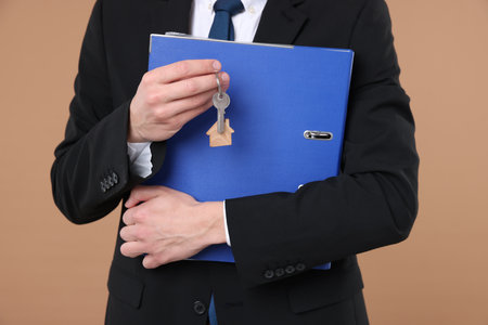 Real estate agent holding key with house shaped keychain and folder on beige background, closeupの写真素材