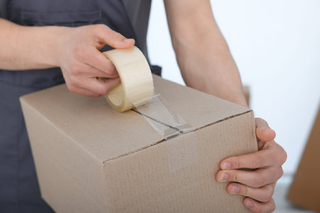 Man sealing cardboard box with adhesive tape indoors, closeup