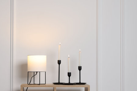 Candlesticks with burning candles and lamp on wooden table near white wall indoors, space for textの写真素材