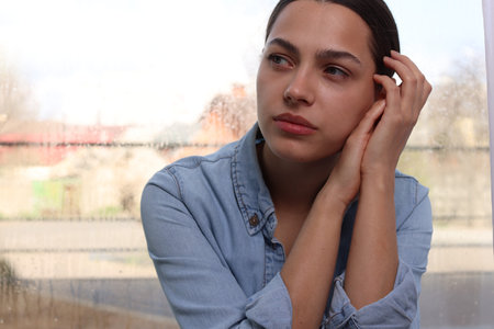 Depressed woman near window at home, space for text