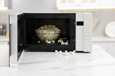 Open microwave oven with bowl of popcorn on white marble table in kitchenの写真素材