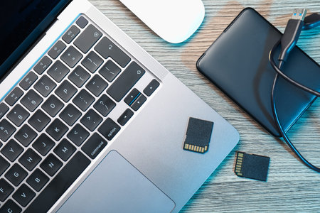 Hard disk drive with USB cable, SD memory cards and laptop on wooden table, flat lay