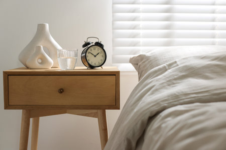 Bedside table with alarm clock, glass of water and vases near bed indoors