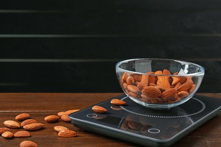 Electronic kitchen scale with bowl of almond nuts on wooden table against black background, closeup. Space for textの写真素材