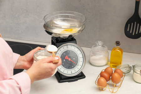 Woman holding spoon with flour near mechanical kitchen scale at white marble countertop indoors, closeupの写真素材