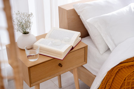 Bedside table with book, flowers in vase, glass of water and glasses near bed indoorsの写真素材