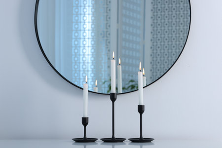 Stylish candlesticks with burning candles on light table near white wall and mirror indoorsの写真素材
