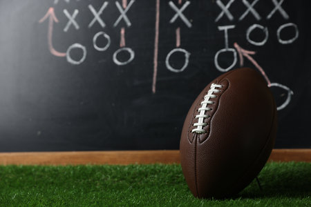 American football ball on green grass against chalkboard with game schemeの写真素材