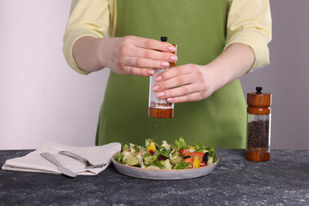 Woman salting tasty salad at dark textured table against white background, closeup