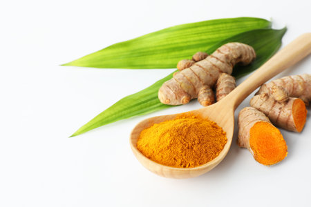 Turmeric powder in spoon, green leaves and roots on white background, closeup. Space for text