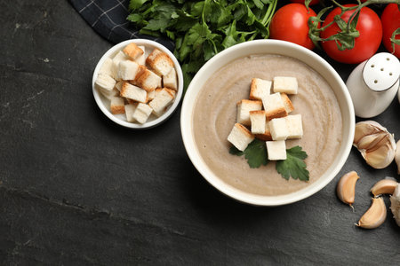 Delicious mushroom cream soup with croutons, parsley and ingredients on black textured table, flat layの写真素材