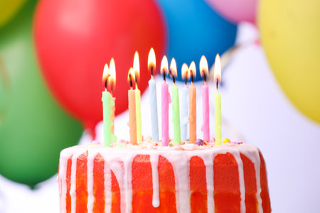 Delicious birthday cake with burning candles and colorful balloons against light background, closeupの写真素材