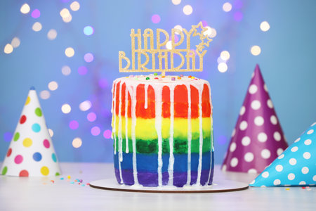 Delicious rainbow birthday cake with topper and party hats on white table against light blue background with color blurred lights, closeupの写真素材