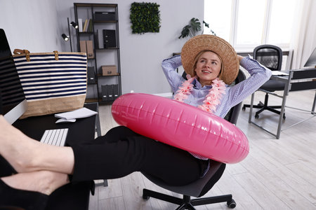 Businesswoman with inflatable ring, flower wreath, bag and straw hat at workplace in officeの写真素材