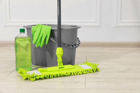 Microfiber mop, bottle of detergent, bucket and gloves on wooden floor near white wall. Space for textの写真素材