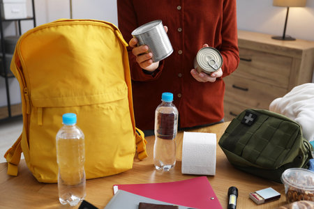 Survival kit. Woman packing different emergency supplies into backpack at wooden table indoors,...