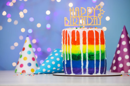 Delicious rainbow birthday cake with topper and party hats on white table against light blue background with color blurred lights, closeupの写真素材