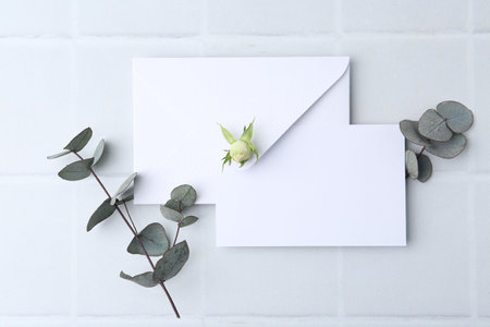 Blank greeting card, paper envelope and eucalyptus branches on light tiled background, flat layの写真素材