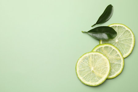 Slices of fresh limes and leaves on pale green background, flat lay. Space for text