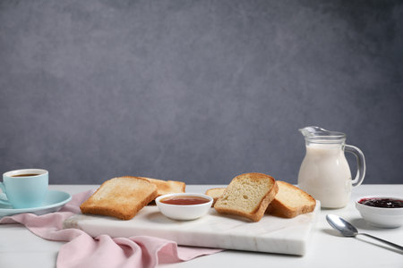 Slices of tasty toasted bread, milk, coffee and jam on white table against grey background. Space...
