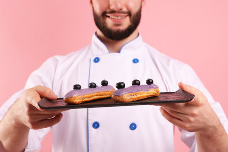 Happy confectioner in uniform holding eclairs on pink background, closeupの写真素材