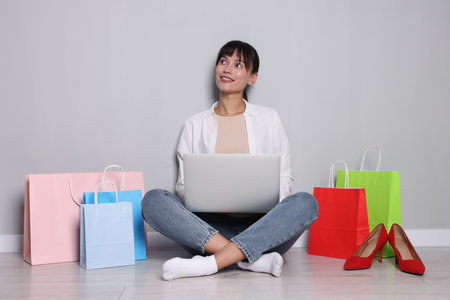 Internet shopping. Happy woman with laptop and colorful bags sitting near gray wallの写真素材