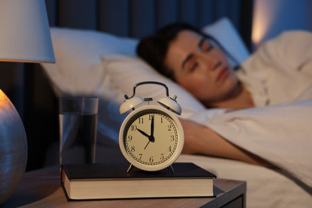 Young woman sleeping in bed at home, focus on alarm clock and book