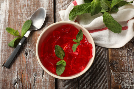 Delicious red jelly cubes in bowl, mint and spoon on wooden table, flat lay