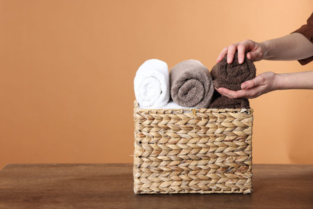 Woman taking clean towel from basket at wooden table against brown background, closeup. Space for textの写真素材