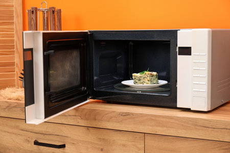 Open microwave oven with piece of lasagna on wooden cabinet indoorsの写真素材