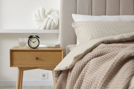 Bedside table with alarm clock, glass of water and book near bed indoors