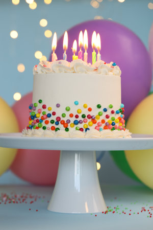 Delicious cake with colorful sprinkles and candles on light blue table against blurred lights, closeupの写真素材