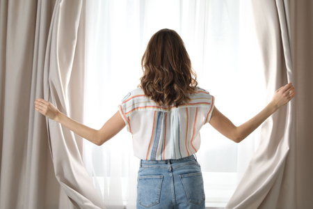 Woman opening window curtains at home in morning, back viewの写真素材