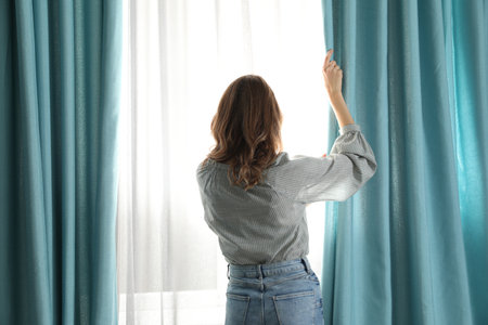 Woman opening window curtains at home in morning, back viewの写真素材