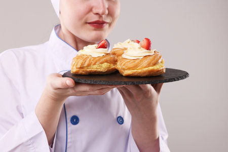 Confectioner in uniform holding delicious profiteroles on light gray background, closeupの写真素材