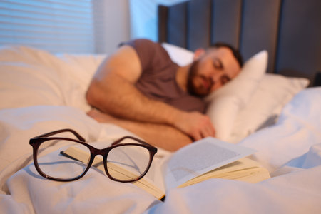 Young man sleeping in bed at home, focus on book and glassesの写真素材