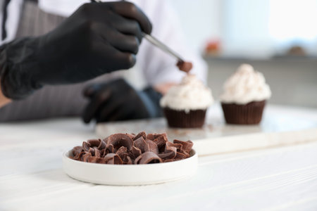 Confectioner decorating tasty cupcake with chocolate shaving at white wooden table in kitchen, selective focusの写真素材