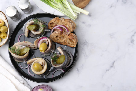 Delicious herring rolls with olives, pickles, dill, onion and bread pieces on light marble table, flat lay. Space for textの写真素材